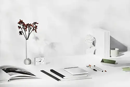 A photograph of a set of notebooks, pens and flowers in a white, minimalist office